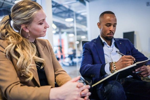 Two meeting and interacting. One is taking notes and the other is analyzing those notes.