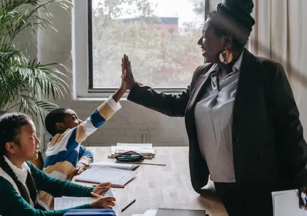 A school administrator gives a high-five to a student during a classroom activity. Another student looks on, creating a positive and encouraging learning environment.