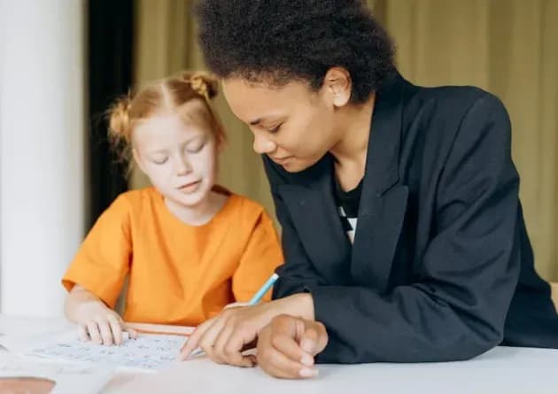 A school counselor assists a young student with her work at a desk. The counselor provides guidance and support, fostering a positive and nurturing learning environment.