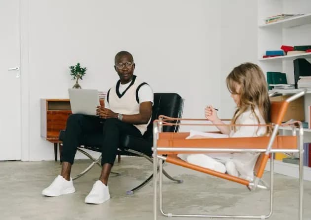 A school psychologist sits with a young student in a modern, minimalist office setting. The psychologist uses a laptop, creating a professional and supportive environment for the student's session.