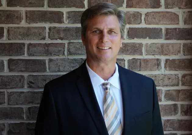 A smiling school vice principal stands in front of a brick wall. He is dressed in a suit and tie, projecting professionalism and confidence.