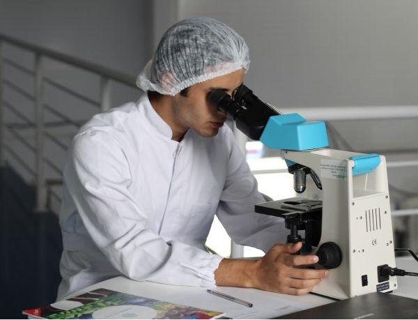 A scientist wearing a lab coat and a hairnet is closely examining a sample through a microscope. The setting is a modern laboratory, with a staircase and lab equipment visible in the background, indicating a professional and research-focused environment.