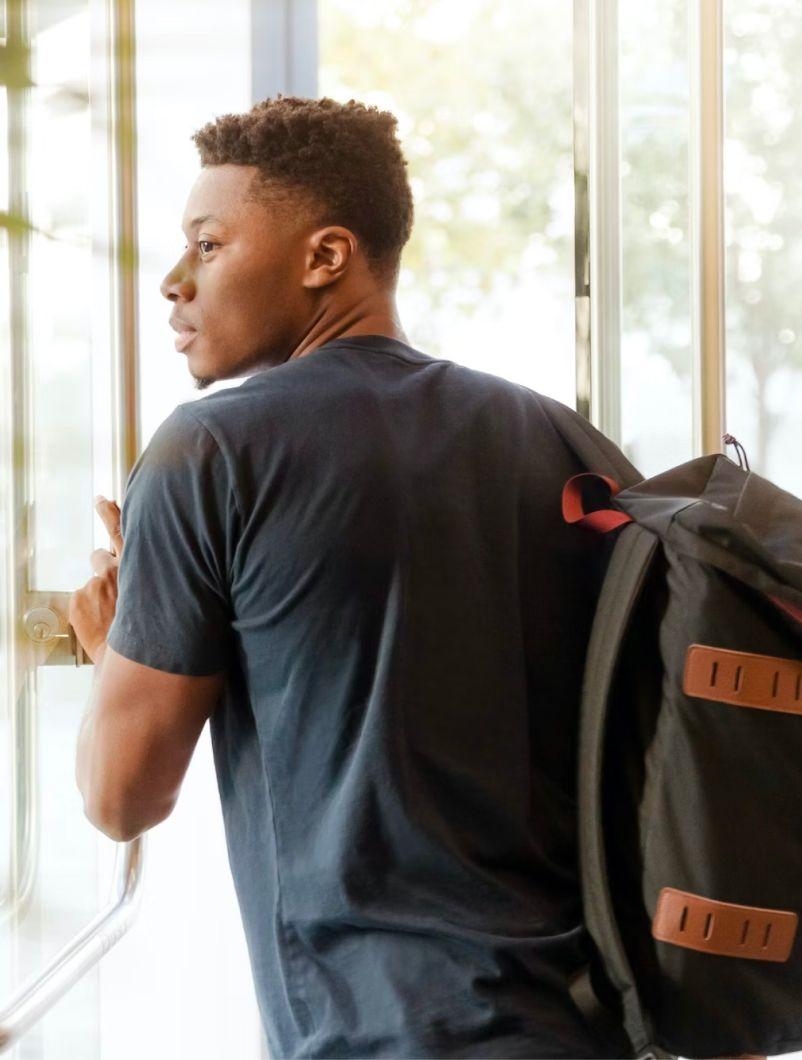 A young man with short curly hair is opening a glass door and looking outside. He is wearing a dark t-shirt and has a black backpack slung over one shoulder. The background shows a bright, sunny day with trees visible through the glass, suggesting he is either entering or leaving a building, ready to take on the day. The scene conveys a sense of anticipation and readiness for the future.