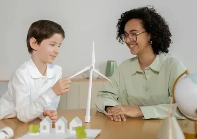 A teacher for individuals with autism smiles as she observes a young boy engaging with a model wind turbine. The scene captures a supportive and interactive educational setting.