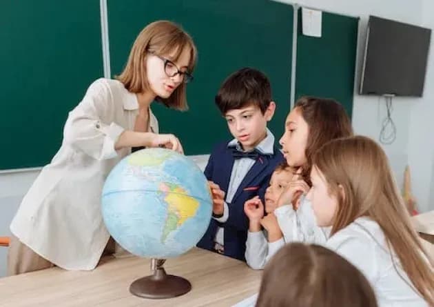 A teacher explains geography to a group of attentive students using a globe in a classroom. The children are gathered around, engaging with the lesson, fostering a collaborative learning environment.