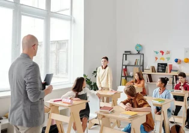 A teacher stands at the front of a classroom, engaging with students who are seated at desks. One student stands to speak, creating an interactive and inclusive learning environment.