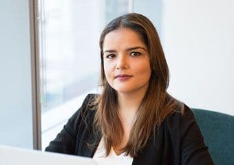 A professional woman with long brown hair is sitting at a desk in an office, looking confidently at the camera. She is dressed in business attire, suggesting her role in a corporate or professional setting.