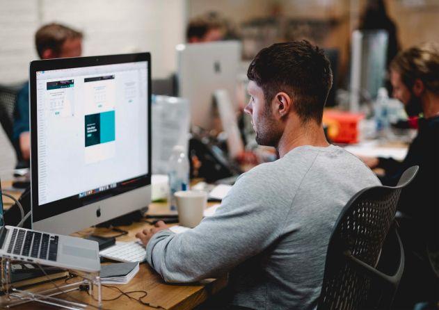 A man is working at a desk, focused on a large computer screen displaying a design application. He is dressed in a light gray shirt, seated in a busy office environment with colleagues working at their desks in the background. The setting suggests a professional workspace, possibly in a design or tech company, indicating a collaborative and productive atmosphere.