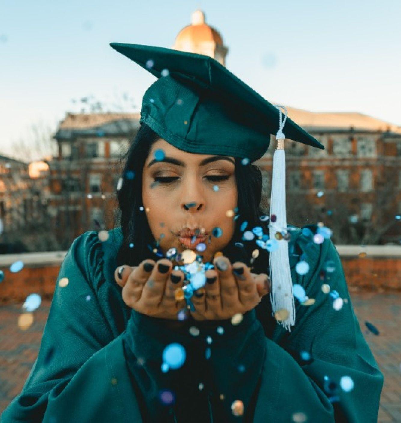 A young woman wearing a green graduation gown and cap is outdoors, blowing confetti from her hands. She is celebrating her graduation with a joyful and festive expression, with a building in the background.