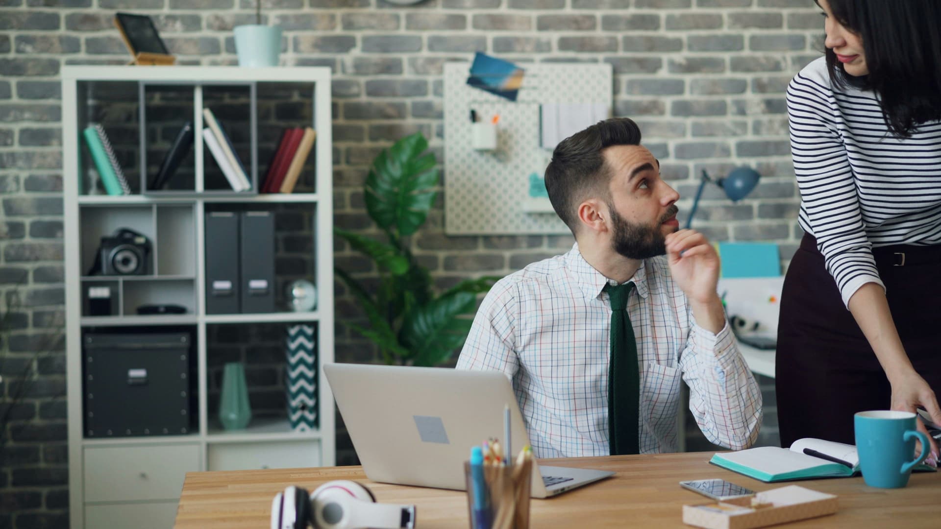 Man sitting at desk, talking to woman standing beside him.