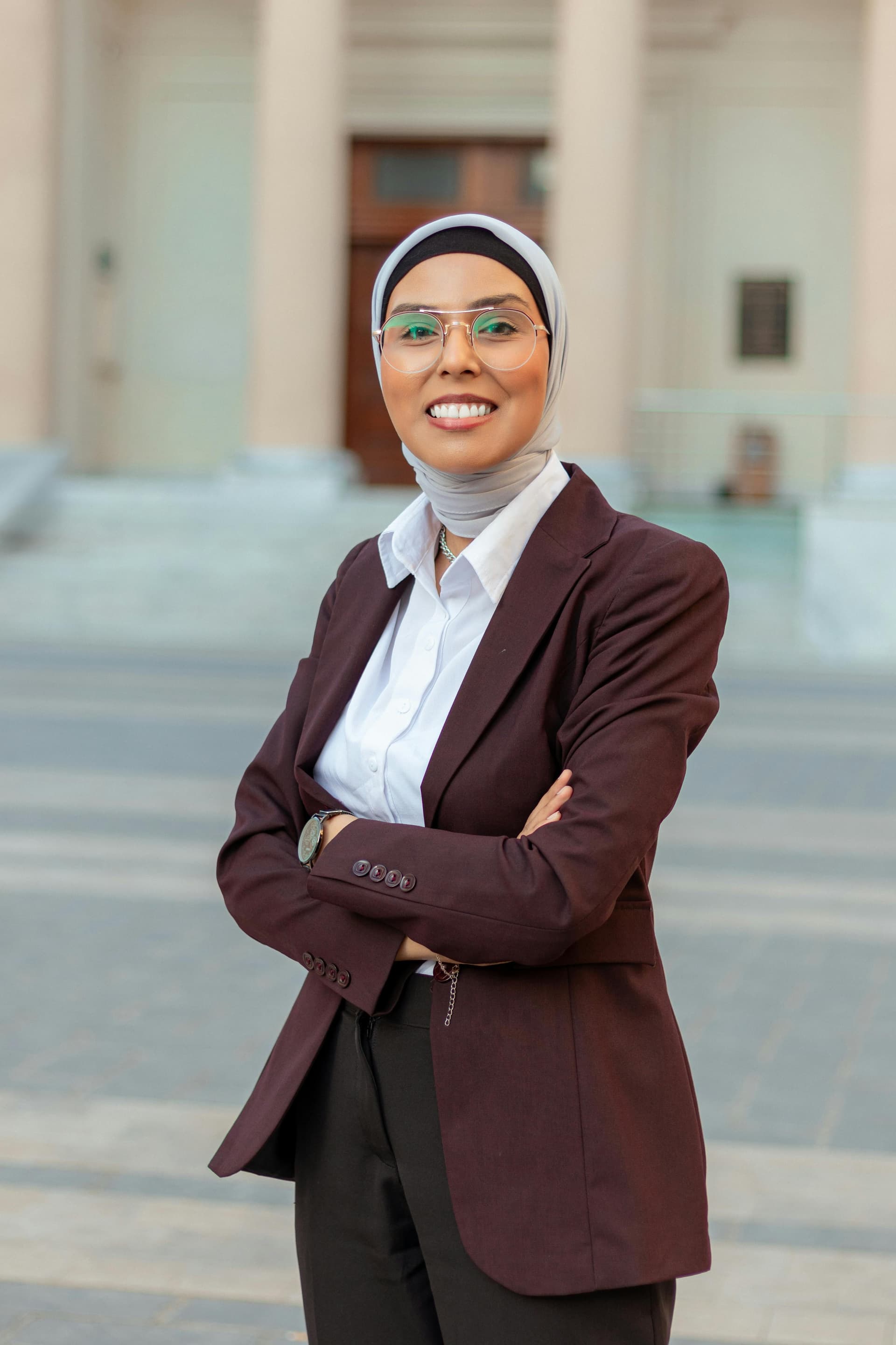 Person in a blazer and hijab smiling with arms crossed.
